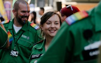 A smiling Advanced First Aider volunteer with glitter on their face and surrounded by other volunteers with rainbow epaulettes. 