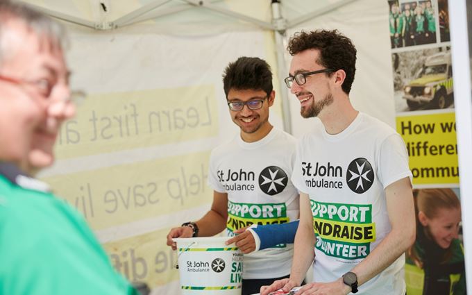 Two fundraising volunteers stood in a tent smiling. One is holding a collection bucket and the other is holding flyers. They're both facing a first aid volunteer.