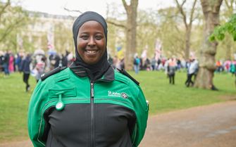 A volunteer standing in a busy park with their arms behind their back and smiling.