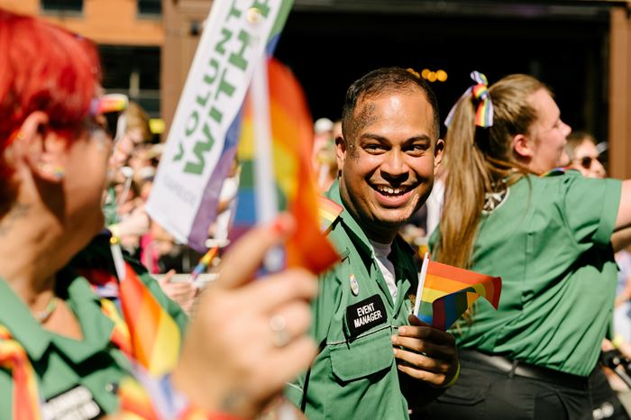A smiling volunteer with an 'Event Manager' badge and with glitter on their face, holding a small rainbow Pride flag. They are surrounded by other volunteer also holding Pride flags.
