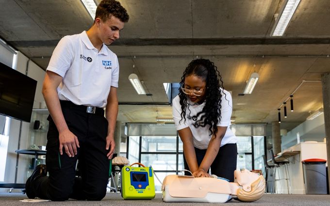 Two teenagers wearing an NHS Cadet polo shirt kneeling on the floor. One of them doing chest compressions on a mannequin with a defibrillator attached.